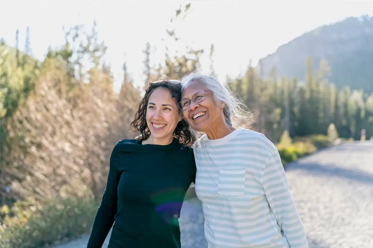 A beautiful mixed race young adult woman embraces her vibrant retirement age mother. The mother and daughter are enjoying a relaxing walk in nature on a beautiful, sunny day. In the background is a mountainous evergreen forest bathed in sunlight.