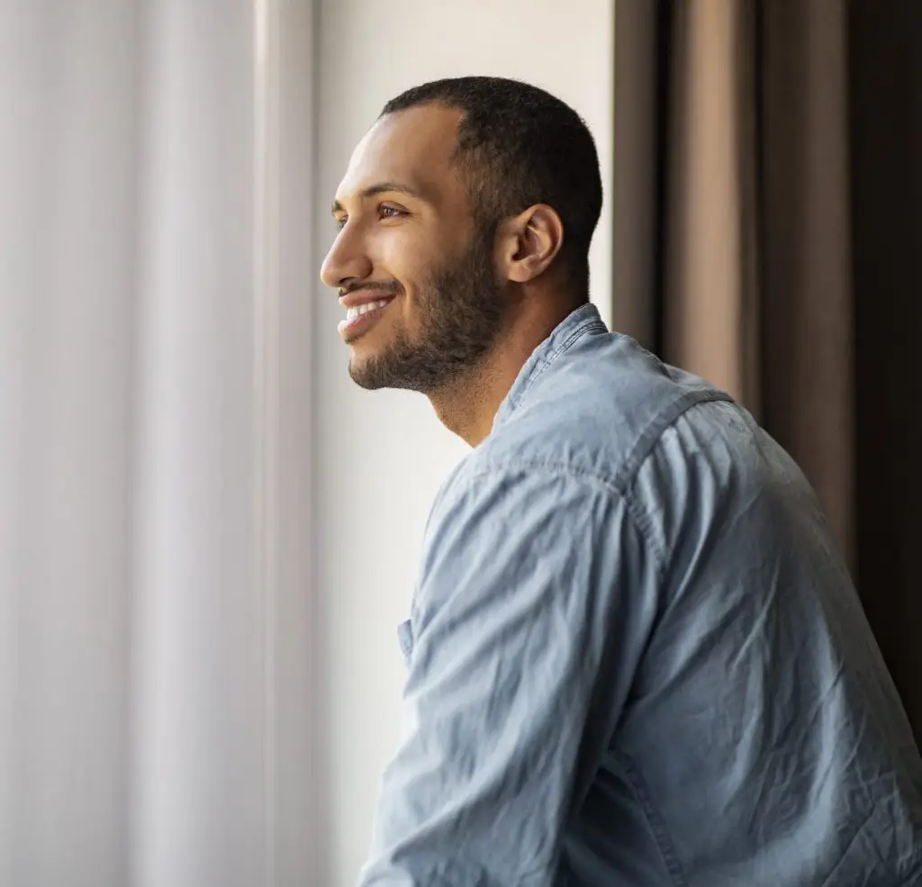 Handsome Smiling African American Guy Standing Near Window At Home And Looking Away, Pensive Young Black Man In Casual Clothes Daydreaming Indoors, Having Good Mood, Side View With Copy Space