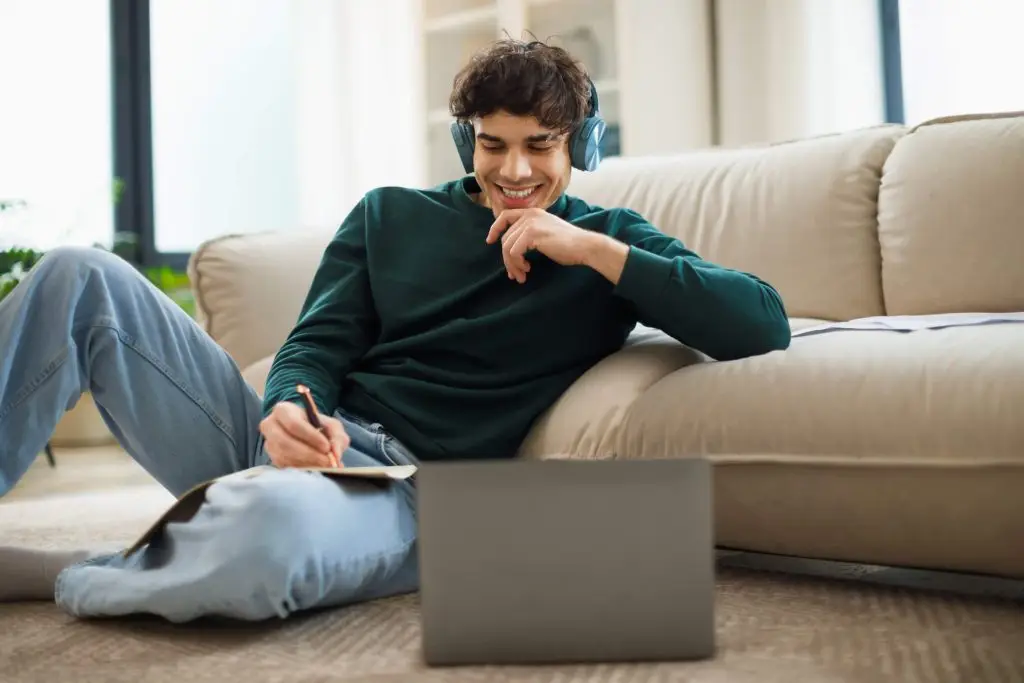 Happy Middle Eastern Student Guy Near Laptop Taking Notes Learning Online Sitting On Floor Using Computer At Home, Wearing Headphones. Modern Distance Education Concept. Selective Focus