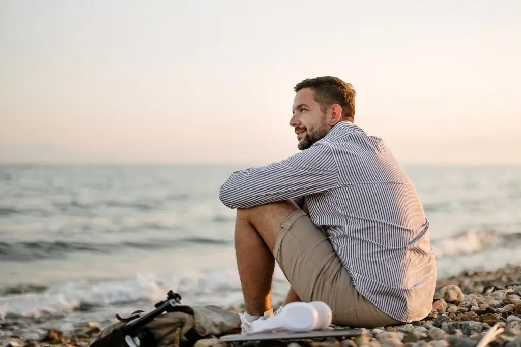 Handsome young man sitting thoughtful on the beach looking in the far
