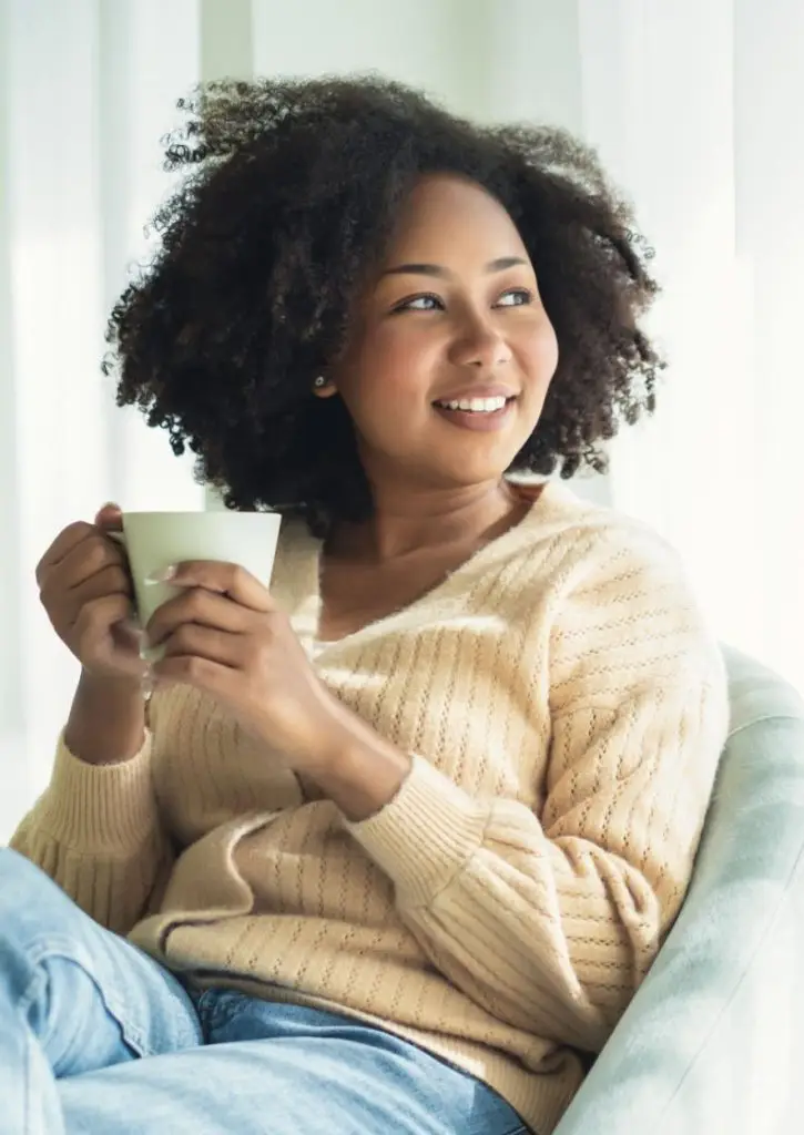 Beautiful young African American woman relaxing on comfortable chair and enjoying a cup of coffee in living room. Smiling calm Black woman enjoy breathing fresh air resting in armchair at home.